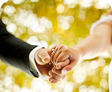 A couple holding hands with wedding rings in a soft-focus outdoor setting.